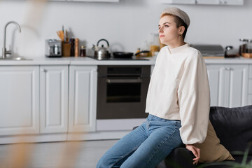 dreamy woman in white pullover sitting in kitchen and looking away