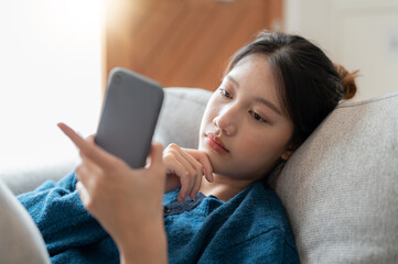 Cheerful young asian woman using mobile phone while sitting on a couch at home.