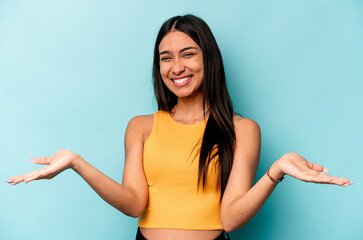 Young hispanic woman isolated on blue background makes scale with arms, feels happy and confident.