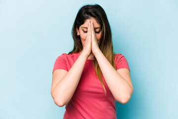 Young caucasian woman isolated on blue background holding hands in pray near mouth, feels confident.