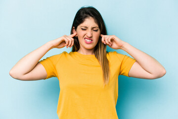 Fototapeta premium Young caucasian woman isolated on blue background covering ears with fingers, stressed and desperate by a loudly ambient.