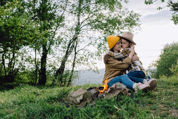 Naklejka na ściany i meble Happy loving young mom and cute little daughter child in hat sit near bonfire on background green forest. Smiling small kid hugs mother, enjoy summer nature. Camping holiday with kids in summer nature
