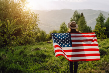 Back view of patriot young woman in hat standing on background summer mountains during beautiful amazing sunset or sunrise, holds proudly usa flag high in air, demonstrates patriotism and freedom