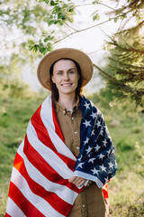 Portrait of calm beautiful young caucasian woman 20s in beige hat wrapped in american flag. Hipster girl smiling and looking at camera on background forest. July 4th forth independence day concept