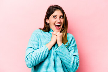 Young caucasian woman isolated on pink background praying for luck, amazed and opening mouth looking to front.