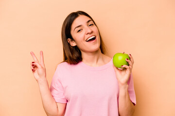 Young hispanic woman holding an apple isolated on beige background joyful and carefree showing a peace symbol with fingers.