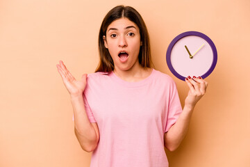 Young hispanic woman holding a clock isolated on beige background surprised and shocked.