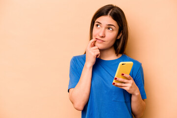 Young caucasian woman holding mobile phone isolated on beige background relaxed thinking about...