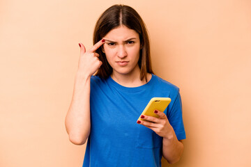 Young caucasian woman holding mobile phone isolated on beige background showing a disappointment gesture with forefinger.