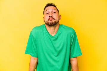 Young hispanic man isolated on yellow background shrugs shoulders and open eyes confused.