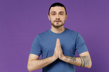 Young smiling man 20s wearing blue t-shirt hold hands folded in prayer gesture, begging about something look camera isolated on plain purple color background studio portrait. People lifestyle concept.