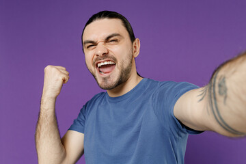 Close up young fun happy man 20s wearing basic blue t-shirt doing selfie shot pov on mobile phone do winner gesture isolated on plain purple color background studio portrait. People lifestyle concept.