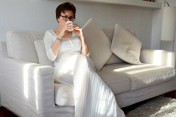 Lifestyle portrait of happy woman in white skirt, shirt relaxing, sitting on couch at cozy home. Drinking coffee. Dreaming