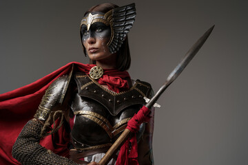 Shot of female barbarian holding spear dressed in steel armor with helmet against grey background.