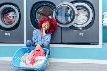 A young woman with red afro hair squatting holding a basket with clothes in a blue automatic laundromat