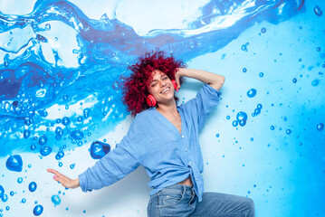 portrait of a happy smiling young latin woman with red afro hair having a good time dancing and listening to music and on her red headphones on a blue background with blue drops