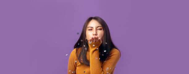 Beautiful young Asian woman cheerful blowing confetti on banner isolated purple studio background. Asian female happy in anniversary concept.