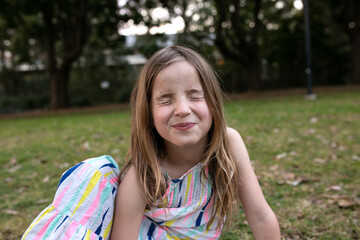 Girl pulling silly face with eyes closed sitting outside on grass