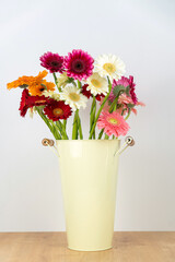 Close-up of a lot of colorful gerberas in a tin vase with handles on a wooden table on a neutral background. Vertical photo