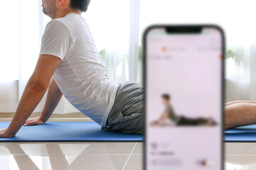 Home exercise using fitness app concept, young man works out, performing a cobra stretch on blue yoga mat, following fitness app, white curtain in background covering houseplants, selective focus