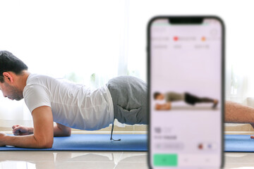 Home exercise using fitness app concept, young man works out, doing forearm plank hold on blue yoga mat, following fitness app, white curtain in background covering houseplants, selective focus