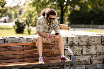 A young man is sitting on a park bench in a public park. He is enjoying the sun and listening to music.