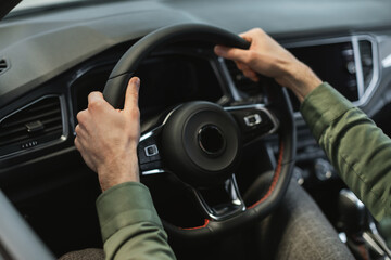 Closeup view of young man holding steering wheel of modern luxury car, going on test drive at modern auto dealership