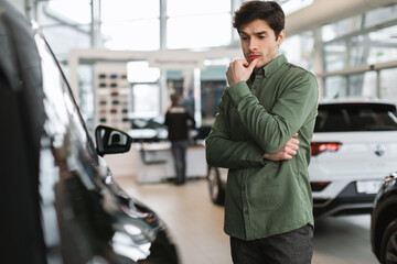 Pensive young guy thinking, making choice, having doubt about buying new car at dealership centre,...