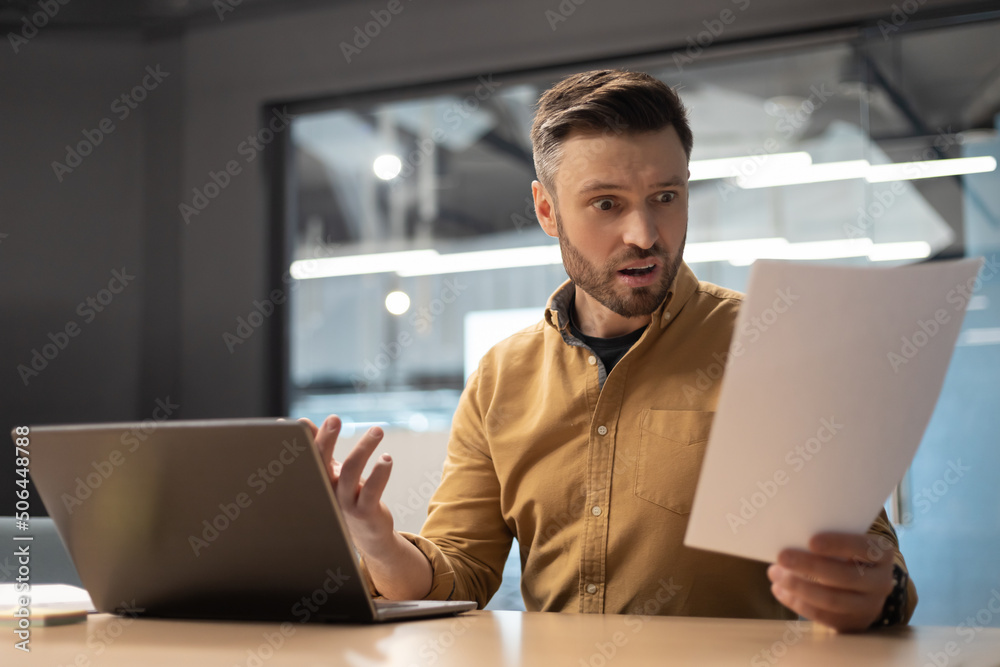 Poster shocked businessman reading papers having problem using laptop in office
