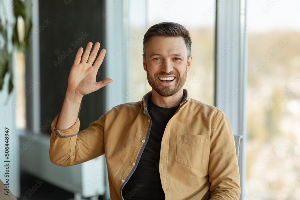 Poster joyful businessman waving hello smiling to camera sitting in office