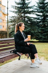 Charming positive young woman in stylish coat resting on bench during coffee break outdoors. Side view of smiling pretty lady holding disposable coffee cup and looking away in park