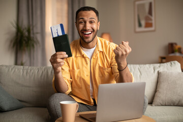 Excited young man with passport and ticket gesturing YES, using laptop to buy hot tour at web travel agency from home - obrazy, fototapety, plakaty