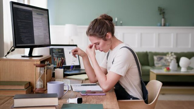 young tired woman sits at desk at home, remote works on computer, stops typing on keyboard, takes off glasses and closes irritated eyes to have a break and rest. Feels overworked and emotional stress.