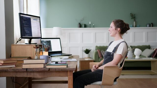 young woman remote works or distance studies in home office sitting at table with computer laptop. gets excited of great news, smiles and laughs, gestures emotionally and waves hands, screams yeah