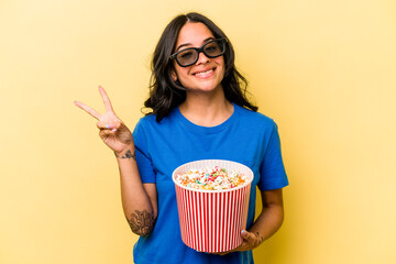 Young hispanic woman holding popcorn isolated on yellow background joyful and carefree showing a peace symbol with fingers.
