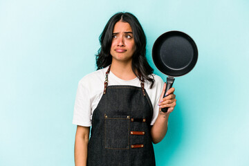 Young hispanic cooker woman holding frying pan isolated on blue background confused, feels doubtful...