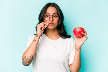 Young hispanic woman holding an apple isolated on blue background with fingers on lips keeping a secret.
