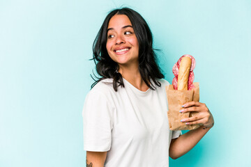 Young hispanic woman eating a sandwich isolated on blue background looks aside smiling, cheerful...