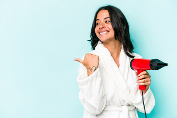 Young hispanic woman holding dryer isolated on blue background points with thumb finger away, laughing and carefree.