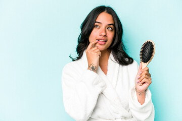 Young hispanic woman holding hairbrush isolated on blue background relaxed thinking about something looking at a copy space.