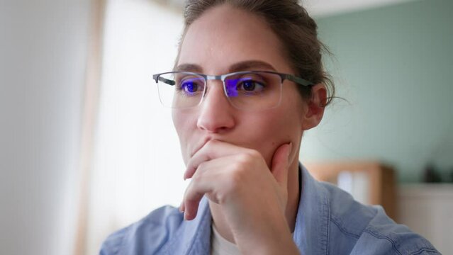 close up front view of young woman in glasses looking in laptop screen, reads and types on keyboard. remote works or studies at home. feels worried and displeased, touches face and breathes out