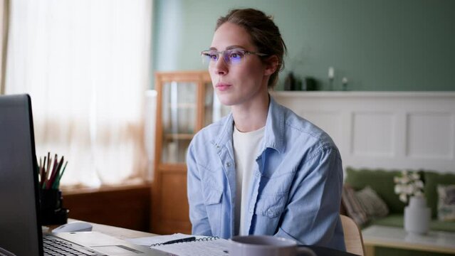 young woman in glasses works at home office on laptop. student passes exam on distant, sits at table. stops typing on keyboard, starts waiting nervously result, bits lip and swings from side to side