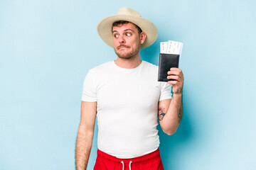 Young caucasian traveler man holding passport isolated on blue background confused, feels doubtful and unsure.