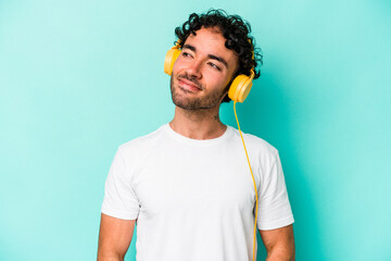 Fototapeta Young caucasian man listening to music isolated on blue background dreaming of achieving goals and purposes obraz