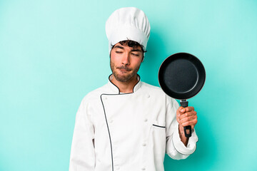 Young caucasian chef man holding flying pan isolated on blue background confused, feels doubtful and unsure.
