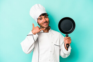 Young caucasian chef man holding flying pan isolated on blue background showing a mobile phone call gesture with fingers.