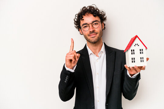 Young caucasian business man holding a toy house isolated on white background showing number one with finger.