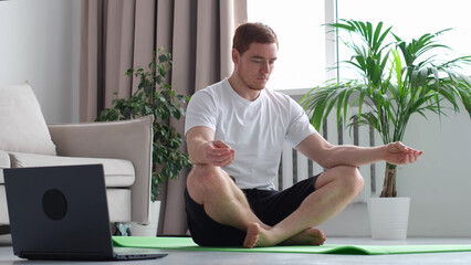 Keep calm concept. Millennial guy meditating with trainer online via laptop connection. Concentrated athletic man meditating after workout at home