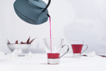 Verter te rojo de bayas en una taza transparente con tetera de hierro azul sobre una mesa blanca y un fondo gris claro	