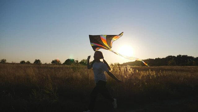 Happy small girl jogging with rainbow kite on rural path at sunset time. Carefree child plays with toy while running through trail near grass field. Cute kid having fun at nature and enjoying freedom